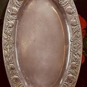 May include: Oval silver-toned serving tray with an embossed apple and leaf border. The tray has a smooth, reflective surface and a decorative handle at each end. The tray is likely made of metal and is suitable for serving food or displaying items.