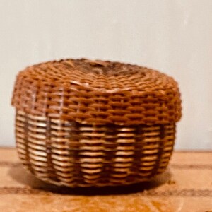 May include: A small, brown wicker basket with a lid. The basket is woven with a light brown and dark brown pattern.