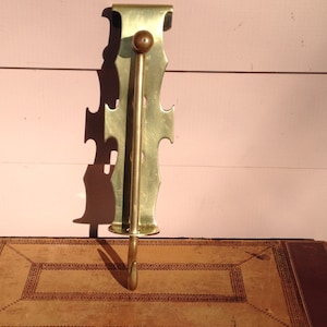 May include: A gold-toned metal coat hook with a decorative design. The hook is mounted on a wall and has a single hook at the bottom.