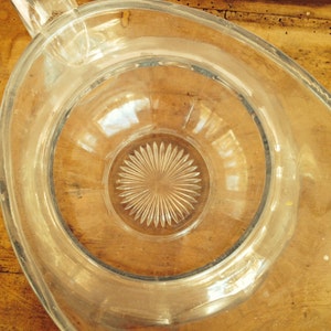May include: A clear glass pitcher with a wide mouth and a textured, starburst-shaped base. The pitcher is empty and sitting on a wooden surface.