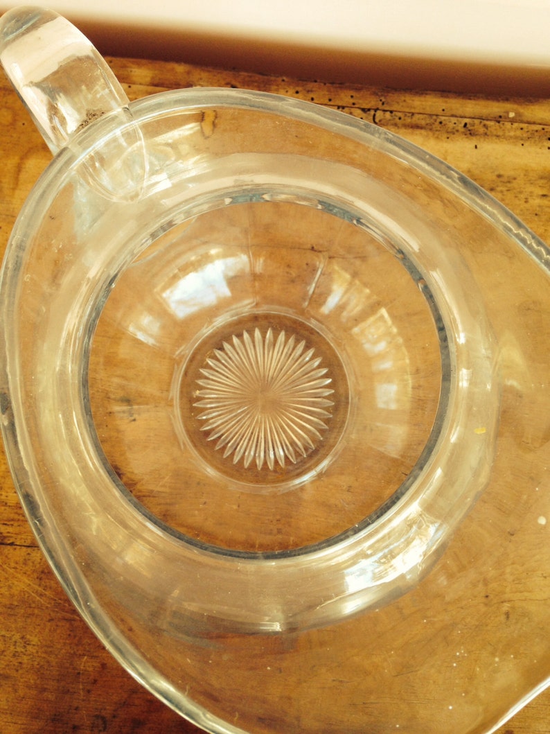 May include: A clear glass pitcher with a wide mouth and a textured, starburst-shaped base. The pitcher is empty and sitting on a wooden surface.