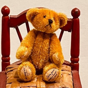 May include: A golden brown teddy bear sitting on a red wooden chair with a woven straw seat. The bear has black eyes and a black nose. The bear's paws have the text "Little Gem Teddy Bear" and "Loving You" written on them.