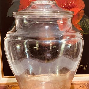 May include: A clear glass apothecary jar with a lid. The jar has a bulbous body and a decorative finial on the lid. The background features a painting with red flowers.