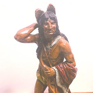 Puede incluir: Estatua detallada de un nativo americano, esculpida con tonos marrones y beige. Lleva un tocado de plumas y una prenda con flecos, con un chal rojo y blanco sobre el brazo. La figura tiene pelo largo y oscuro.
