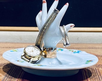 Op de afbeelding: Een witte keramische sieradenhouder in de vorm van een hand met een ronde voet voorzien van kleine blauwe en groene bloemaccenten. Een goudkleurig polshorloge met een textuur metalen band en een zilverkleurige ring sieren de vingers.