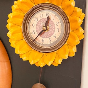 May include: A decorative wall clock shaped like a sunflower. The clock face has a light-colored background with black numerals and hands. The clock is surrounded by bright yellow petals and has a brown pendulum.