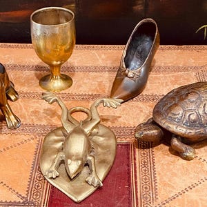 May include: A set of five brass figurines: a dog, a frog on a heart-shaped base, a turtle, a goblet, and a shoe. The figurines are all in a vintage style and have a warm, golden color.