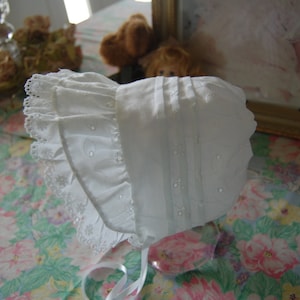 May include: A white lace bonnet with a ruffled edge and embroidered details. The bonnet is made of a soft, delicate fabric and has a simple, elegant design.