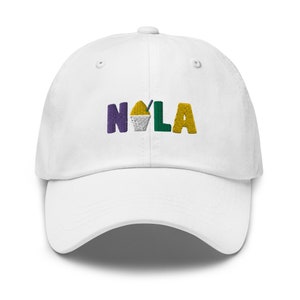 Puede incluir: Gorra de béisbol blanca con un diseño bordado de la palabra "NOLA" en morado, verde y amarillo con un pequeño gráfico de cono de nieve en el medio de la palabra.