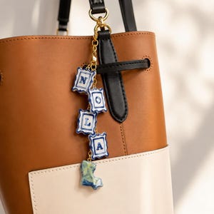 May include: A gold-toned metal chain bag charm featuring four white ceramic rectangular tiles with blue scalloped edges and blue letters spelling NOLA, finished with a small, irregular, light blue and green ceramic shape at the bottom.