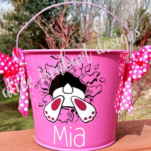 Puede incluir: Cesta de Pascua de metal rosa con un conejo blanco asomándose por un agujero. La cesta tiene una cinta de lunares blancos y el nombre "Mia" está impreso en la parte delantera.