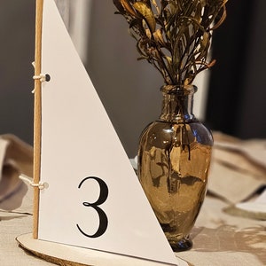 May include: A wooden sailboat table number with a white sail and the number "3" printed on it. The sailboat is sitting on a wooden slice and is next to a vase with dried flowers.