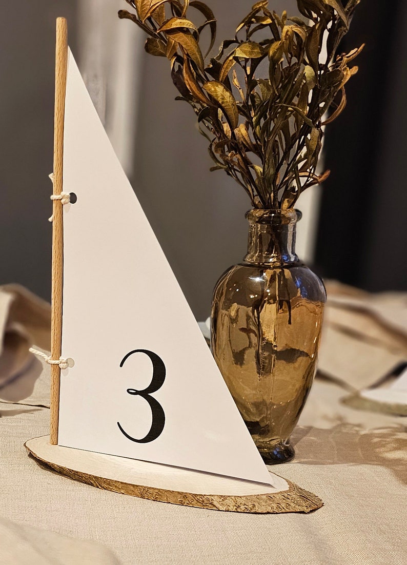 May include: A wooden sailboat table number with a white sail and the number "3" printed on it. The sailboat is sitting on a wooden slice and is next to a vase with dried flowers.