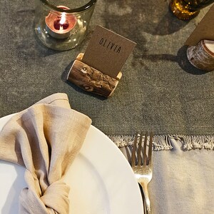 May include: A rustic wooden table number holder with a brown card that says "Table" on it. A second holder has a brown card that says "Olivia". A white plate with a beige linen napkin is on the table. A fork is on the right side of the plate.