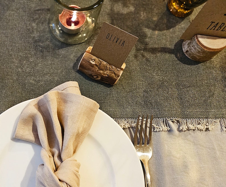May include: A rustic wooden table number holder with a brown card that says "Table" on it. A second holder has a brown card that says "Olivia". A white plate with a beige linen napkin is on the table. A fork is on the right side of the plate.