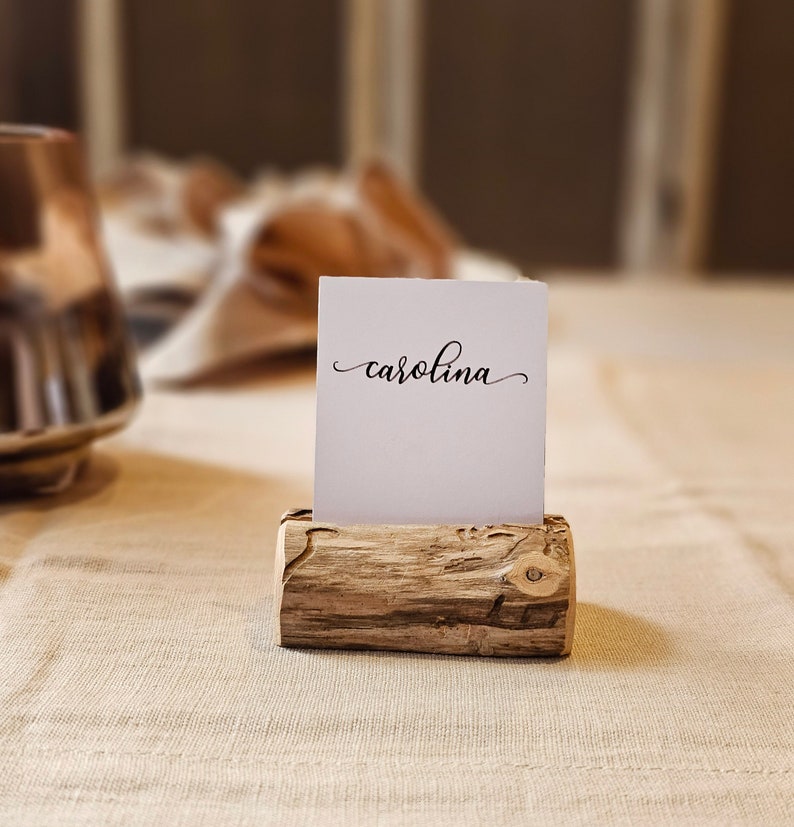 May include: A white card with the name "carolina" written in cursive sits in a wooden place card holder. The holder is made from a natural piece of wood with a rough texture.