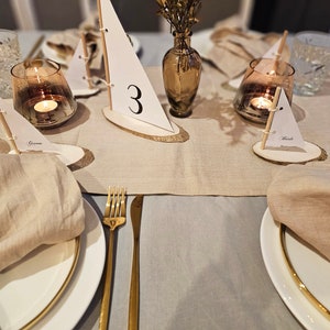 May include: A beige linen tablecloth with gold flatware and white plates. There are small wooden sailboats with numbers and names on them, used as place cards. The sailboats are on small wooden rounds. There are also small glass candle holders with candles.