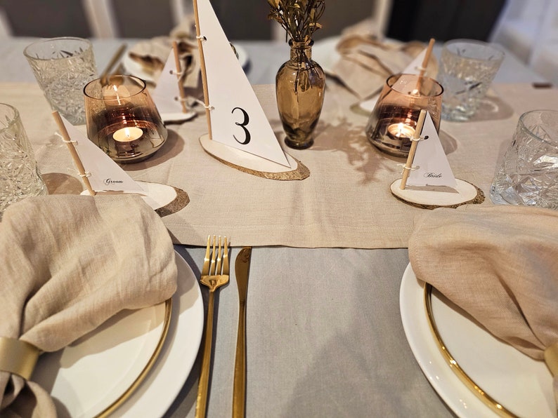 May include: A beige linen tablecloth with gold flatware and white plates. There are small wooden sailboats with numbers and names on them, used as place cards. The sailboats are on small wooden rounds. There are also small glass candle holders with candles.