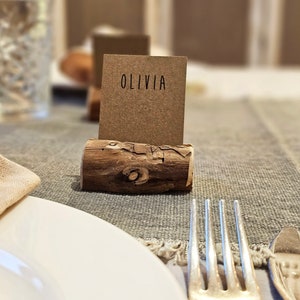 May include: A rustic wooden place card holder with a brown card that says "Olivia" on it. The place card is on a gray linen tablecloth with a white plate and silver fork.
