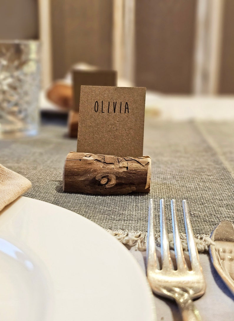 May include: A rustic wooden place card holder with a brown card that says "Olivia" on it. The place card is on a gray linen tablecloth with a white plate and silver fork.
