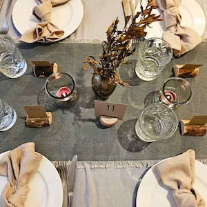 May include: A table setting with a gray linen tablecloth, white plates, and brown napkins. There are clear glasses, candles, and wooden place card holders with the text "Olivia" and "Table 11".