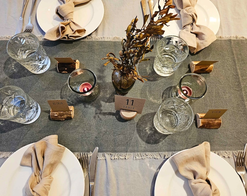 May include: A table setting with a gray linen tablecloth, white plates, and brown napkins. There are clear glasses, candles, and wooden place card holders with the text "Olivia" and "Table 11".