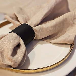 Könnte beinhalten: Eine beige Leinen-Serviette mit einem schwarzen Serviettenring auf einem weißen Teller mit goldfarbenem Rand.