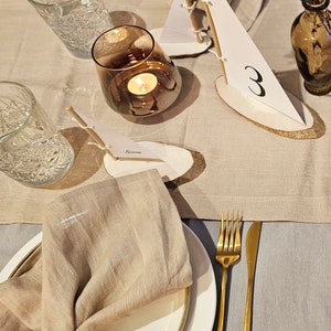 May include: A beige linen napkin with a gold napkin ring is placed on a white plate with a gold rim. A gold fork and knife are on a beige linen tablecloth. A small wooden sailboat with a white sail and the number "3" written on it is on the table. A clear glass with a candle inside is on the table.