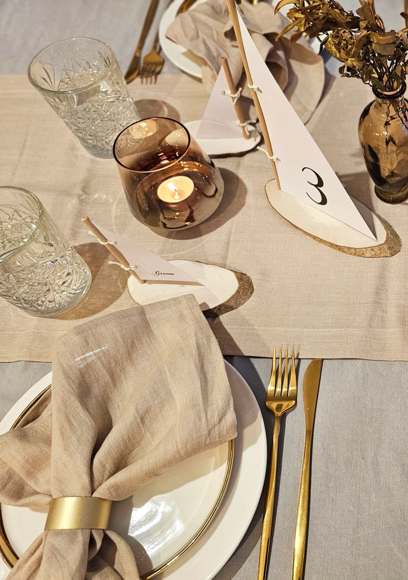 May include: A beige linen napkin with a gold napkin ring is placed on a white plate with a gold rim. A gold fork and knife are on a beige linen tablecloth. A small wooden sailboat with a white sail and the number "3" written on it is on the table. A clear glass with a candle inside is on the table.