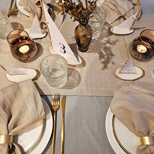 May include: A table setting with a beige linen tablecloth and place settings featuring white plates, gold flatware, and beige linen napkins with gold napkin rings. Each place setting includes a small wooden slice with a paper sailboat and a number. A vase with dried flowers and a candle in a glass holder are also on the table.
