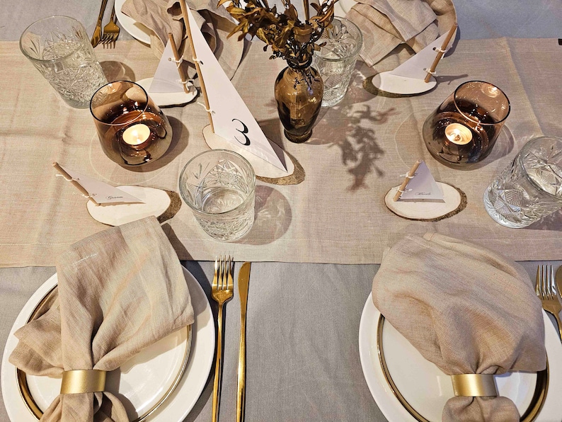 May include: A table setting with a beige linen tablecloth and place settings featuring white plates, gold flatware, and beige linen napkins with gold napkin rings. Each place setting includes a small wooden slice with a paper sailboat and a number. A vase with dried flowers and a candle in a glass holder are also on the table.