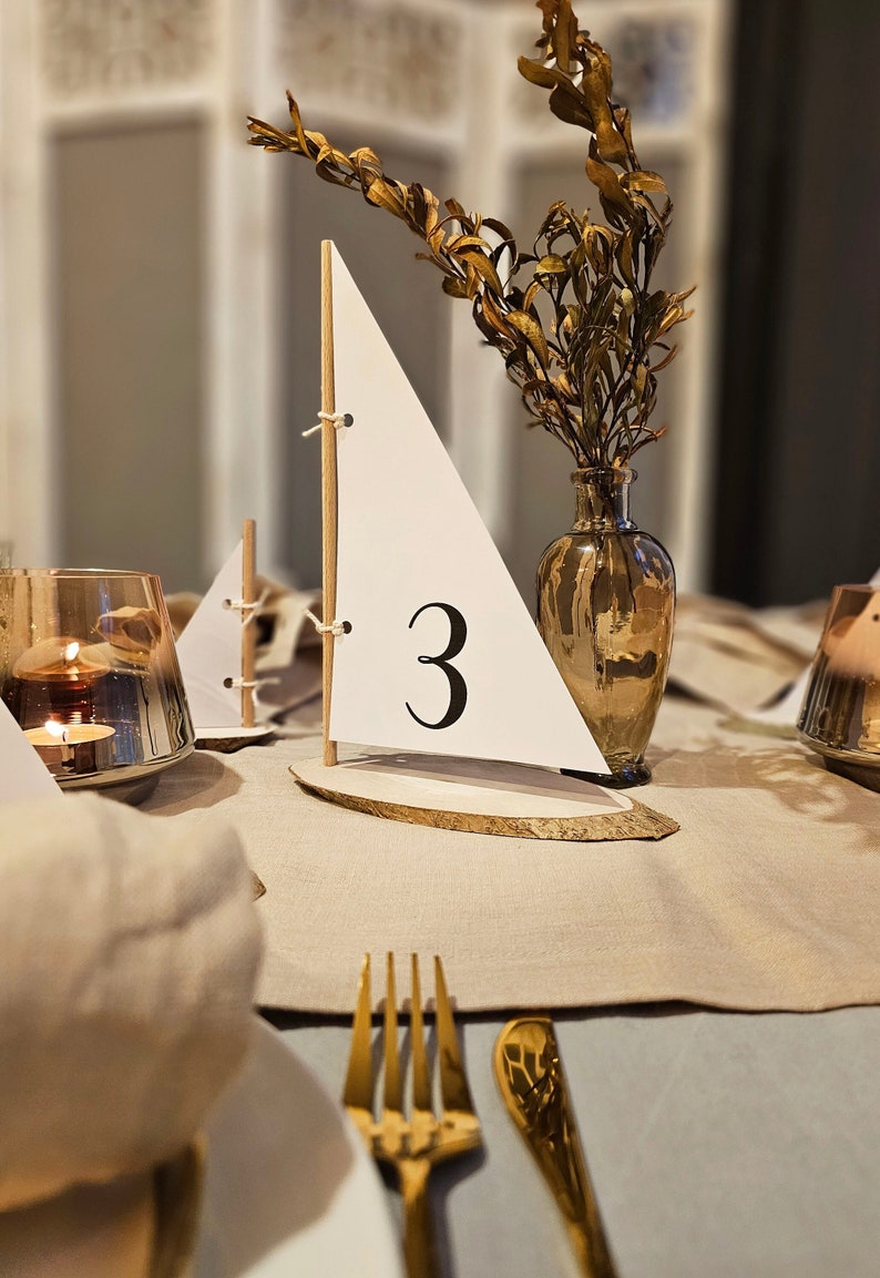 May include: A white sailboat table number with the number "3" on it. The sailboat is on a wooden base and is next to a vase with dried flowers. The table is set with a gold fork and knife.