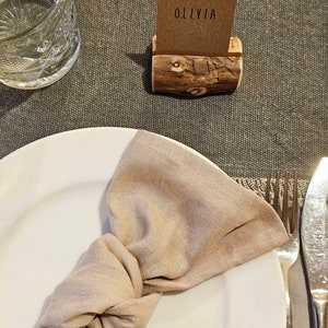 May include: A white plate with a beige linen napkin tied in a knot. A wooden name card holder with the name "Olivia" on a brown card sits on a brown table.
