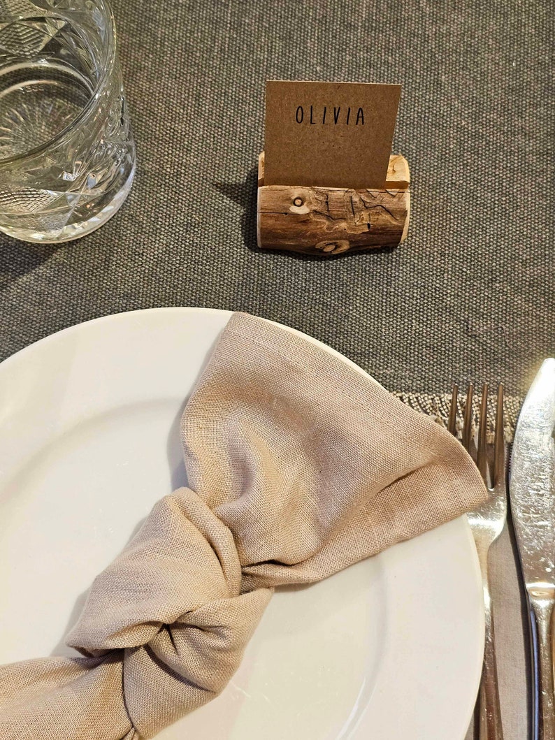 May include: A white plate with a beige linen napkin tied in a knot. A wooden name card holder with the name "Olivia" on a brown card sits on a brown table.
