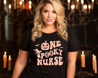 May include: Black t-shirt with the text "One Spooky Nurse" in a pink, retro font, featuring a jack-o'-lantern and bats. The shirt is paired with dark blue leggings. The scene is set with lit candles and Halloween decorations.