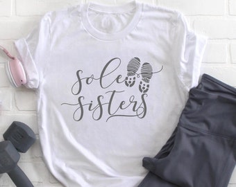 May include: White t-shirt with the text "Sole Sisters" in black cursive lettering and a graphic of two footprints. The shirt is laid out on a white brick background with a pair of grey leggings, a pink pair of headphones, and a grey dumbbell.
