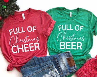 Puede incluir: Dos camisetas con temática navideña, una roja con el texto "Full of Christmas Cheer" y otra verde con el texto "Full of Christmas Beer". Las camisetas están dobladas y se muestran con un par de pantalones vaqueros azules y una boina de punto roja.