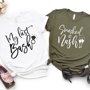 May include: Two t-shirts, one white and one olive green, with text and graphics. The white shirt says "My last Bash" with a diamond ring graphic. The olive green shirt says "Smashed in Nash" with champagne glasses. Accessories include a bottle of pink champagne and black heels.