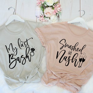 May include: Two t-shirts on white hangers. One is light gray with the text "My last Bash" and a diamond ring graphic. The other is light pink with the text "Smashed in Nash" and champagne glasses.