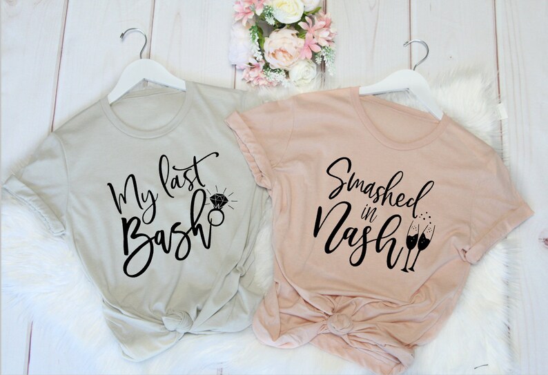 May include: Two t-shirts on white hangers. One is light gray with the text "My last Bash" and a diamond ring graphic. The other is light pink with the text "Smashed in Nash" and champagne glasses.