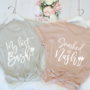 May include: Two t-shirts on white hangers. One is light gray with the text "My last Bash" and a diamond ring graphic. The other is blush pink with "Smashed in Nash" and champagne glasses.