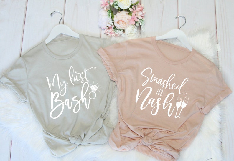 May include: Two t-shirts on white hangers. One is light gray with the text "My last Bash" and a diamond ring graphic. The other is blush pink with "Smashed in Nash" and champagne glasses.