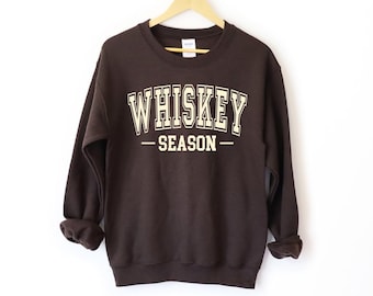 May include: A dark brown sweatshirt with the words "WHISKEY SEASON" printed in a cream-colored, collegiate-style font. The sweatshirt is hanging on a wooden hanger against a white background.