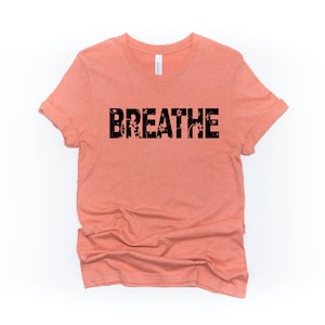 May include: A peach-colored t-shirt with the word "BREATHE" printed in black letters. The letters are filled with a floral pattern.