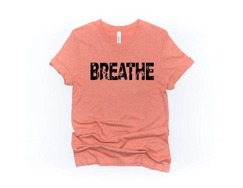 May include: A peach-colored t-shirt with the word "BREATHE" printed in black letters. The letters are filled with a floral pattern.