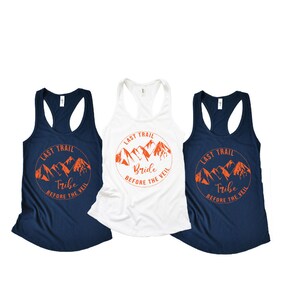May include: Three tank tops with a mountain range graphic and the text "Last Trail" in a circle. The top on the left says "Tribe" and the top in the middle says "Bride". The top on the right says "Tribe".