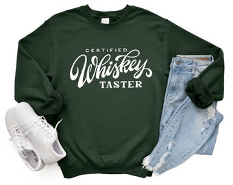 May include: A dark green sweatshirt with the text "CERTIFIED Whiskey TASTER" in white lettering. The sweatshirt is laid flat on a white background with a pair of white sneakers and light blue jeans.