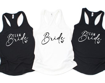 May include: Three tank tops with the word "Bride" written in a script font. One tank top is white with black text, and the other two are black with white text. One of the black tank tops has the word "Team" in front of "Bride".