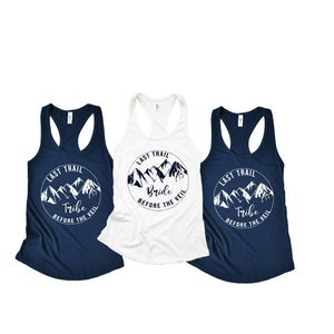 May include: Three white tank tops with a navy blue mountain graphic and the text "Last Trail" and either "Bride", "Tribe", or "Tribe" printed on the front.