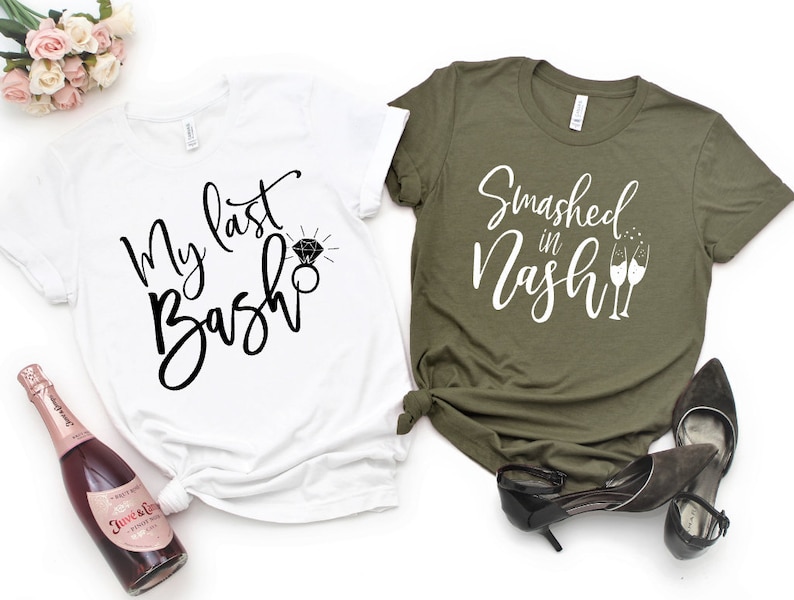 May include: Two t-shirts, one white and one olive green, with text and graphics. The white shirt says "My last Bash" with a diamond ring graphic. The olive green shirt says "Smashed in Nash" with champagne glasses. Accessories include a bottle of pink champagne and black heels.